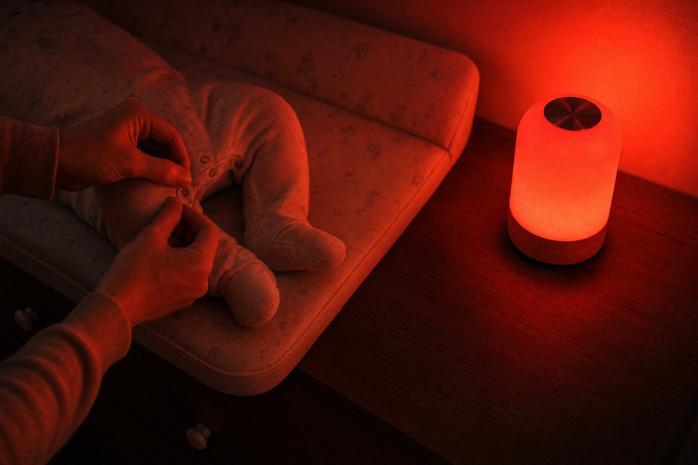 A baby's legs in a sleepsuit and a parent's hands doing up the poppers, lit only by the red glow of the Driftback lamp