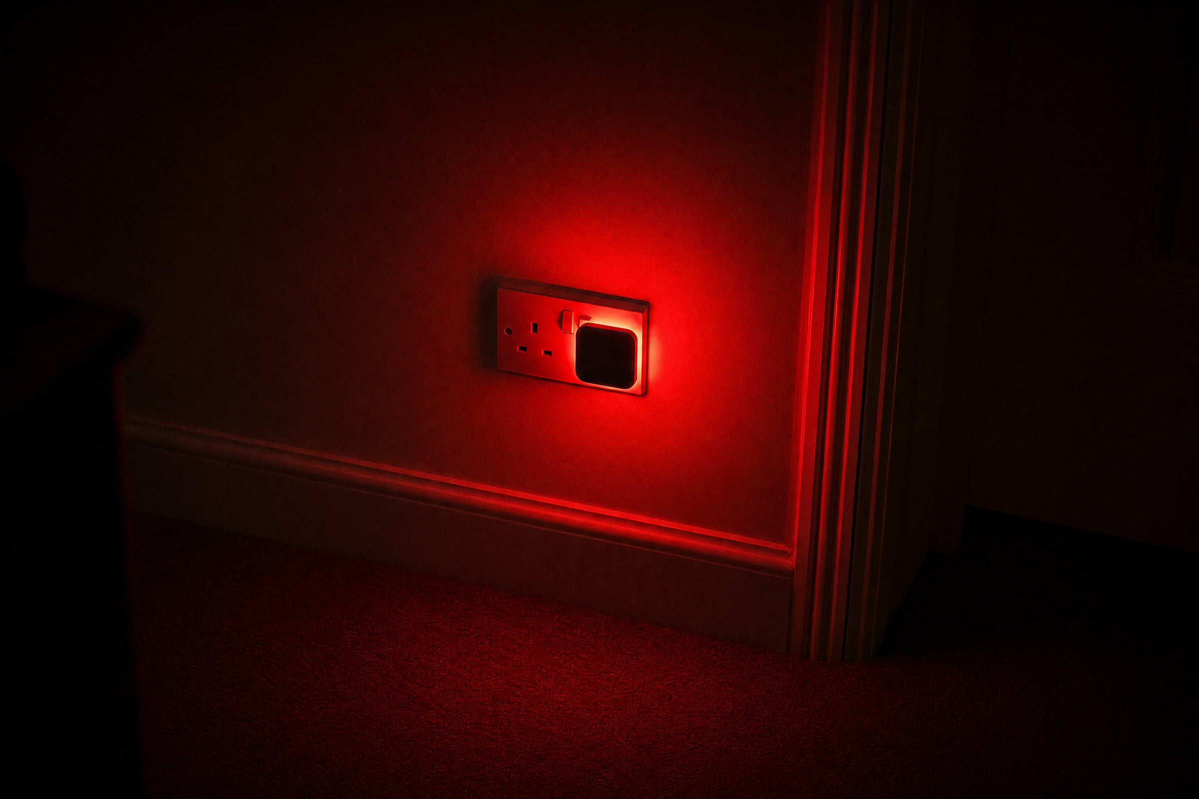 A red night light plugged into a UK wall socket in a dark hallway, glowing softly onto the skirting board.